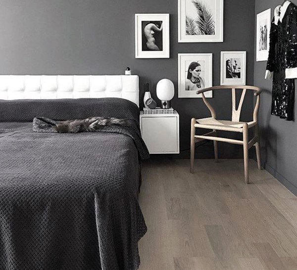 Modern bedroom with a grey bed, white headboard, wooden chair, and framed photos on a dark wall. Elegant decor