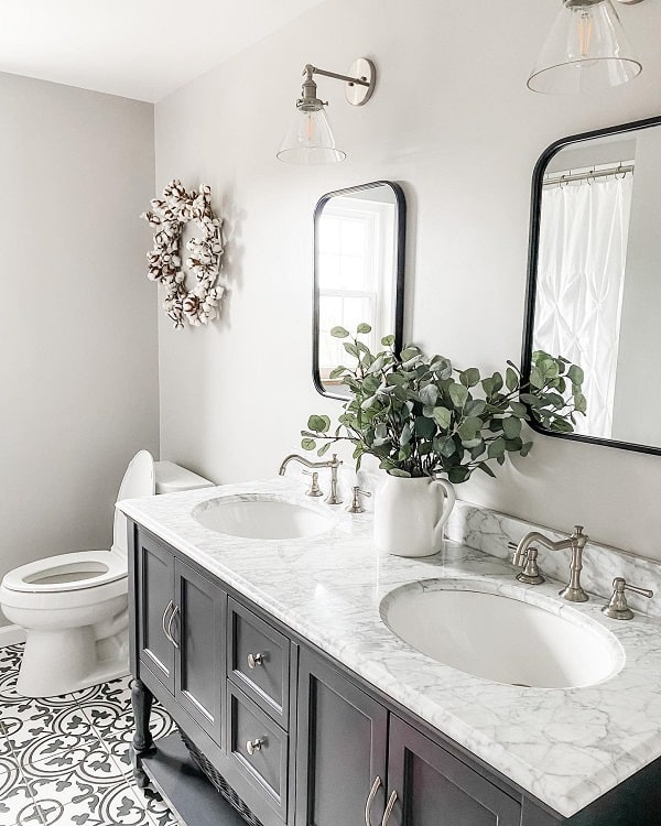 Elegant farmhouse bathroom with dual sinks, marble countertop, greenery in a vase, and decorative mirrors on a tiled floor