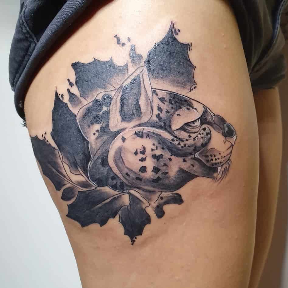 A black and grey tattoo of a jaguar’s face with detailed shading, surrounded by abstract leaves, placed on the upper thigh.