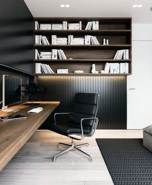 Modern home office with black panel walls, wooden desk, shelves, and swivel chair.