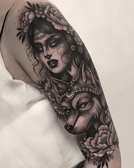 Black and grey tattoo of a mystical gypsy woman with floral details, beaded jewelry, and a regal wolf, covering the upper arm.