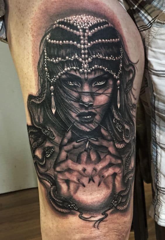 Dark and detailed black and grey tattoo of a mystical woman with piercing eyes, an ornate pearl headdress, and snakes wrapping around her hands.