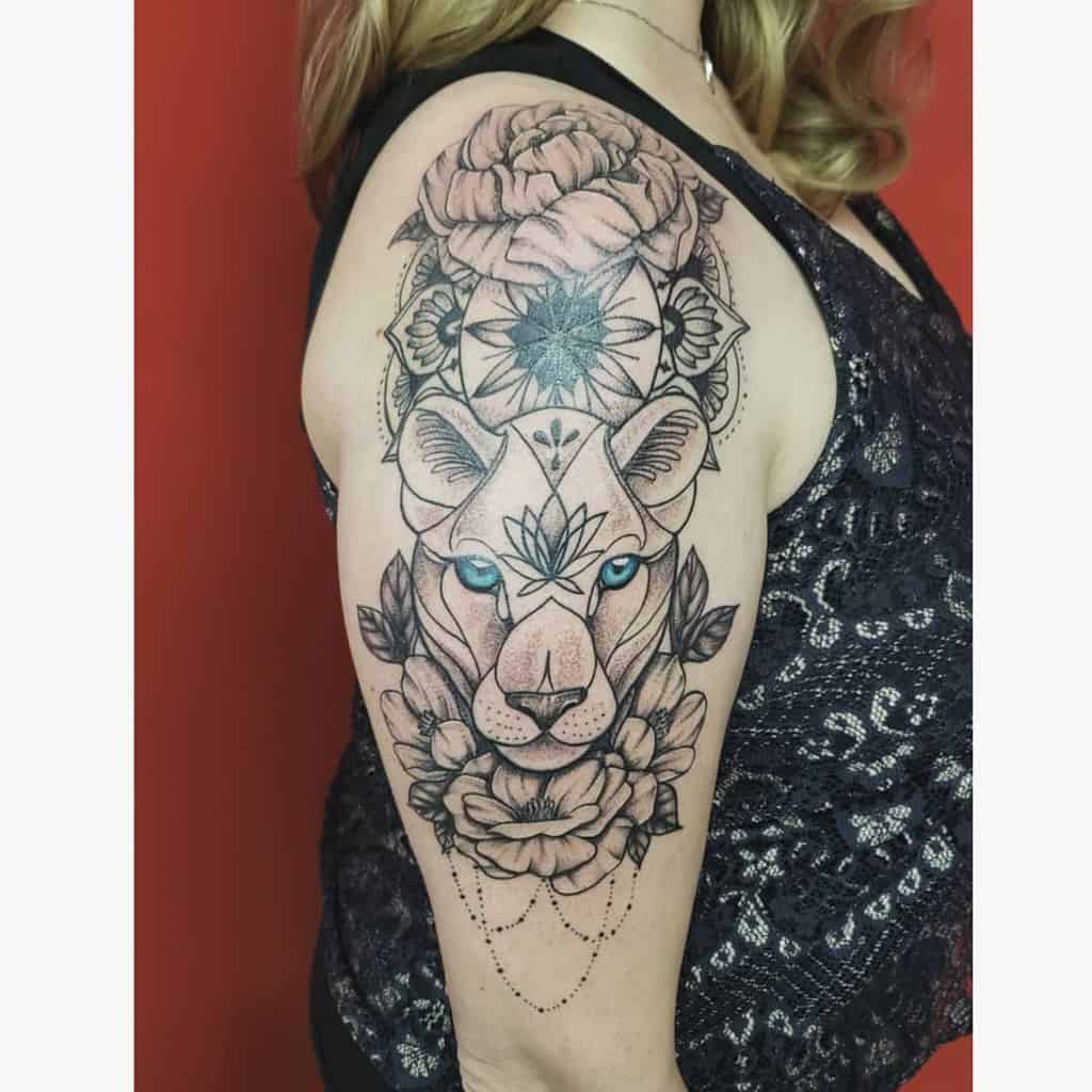 Intricate black and grey lioness tattoo on the upper arm, featuring bold mandala patterns, floral elements, and striking blue eyes.