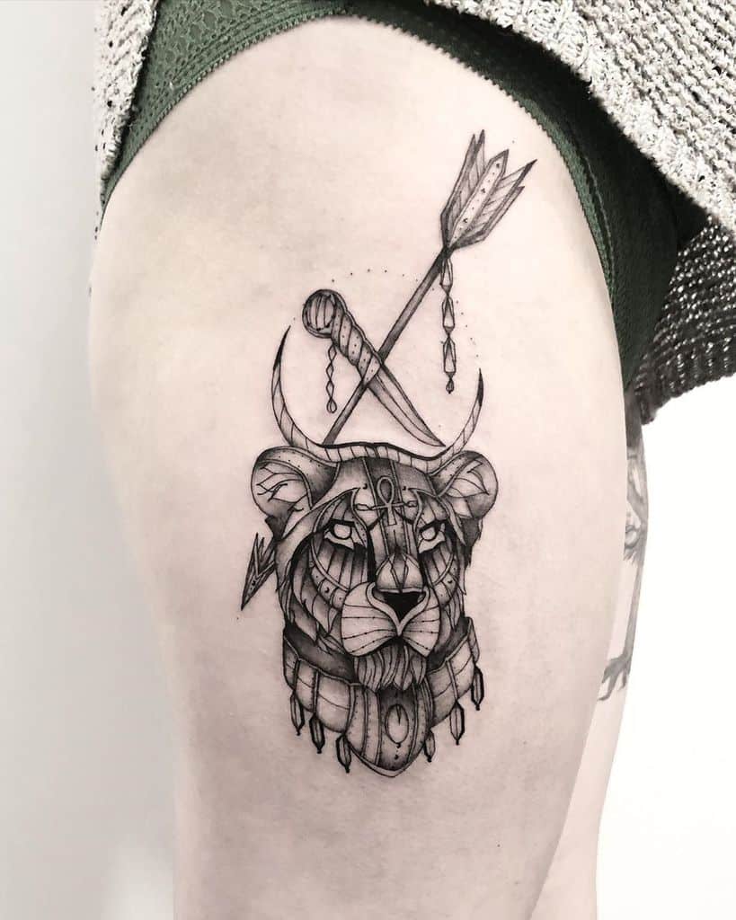 Intricate warrior lioness tattoo on the thigh, featuring tribal-inspired armor, an arrow, and crescent moons for a powerful, mystical look.