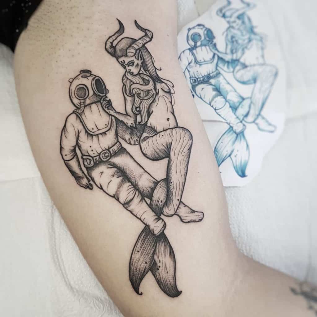 Alt text: "Black and grey tattoo of a horned mermaid entwined with a deep-sea diver in an old-fashioned helmet, creating a surreal and mystical scene.