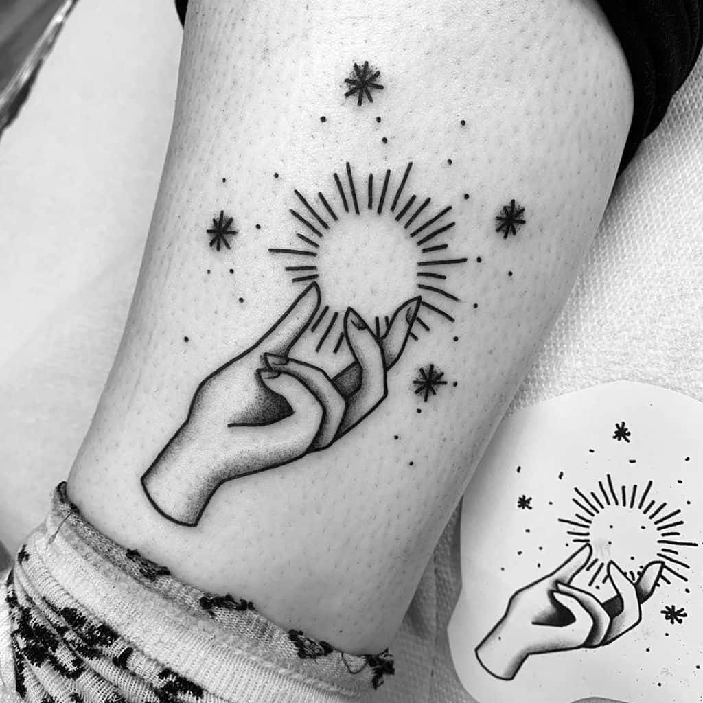 Minimalist black and grey tattoo of two hands holding a radiant sun, surrounded by stars and delicate dot work on the thigh.