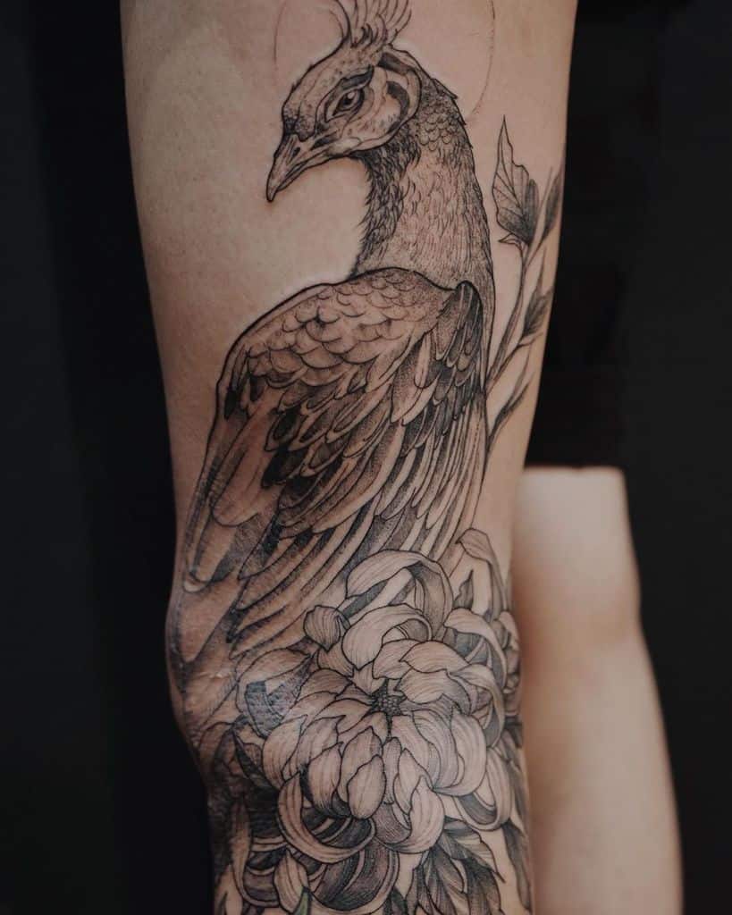 Black and white peacock tattoo with detailed feathers and flowers, featuring a soft circular halo in the background.