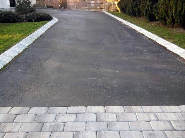 Asphalt driveway with stone edging and a paver threshold near landscaped greenery.