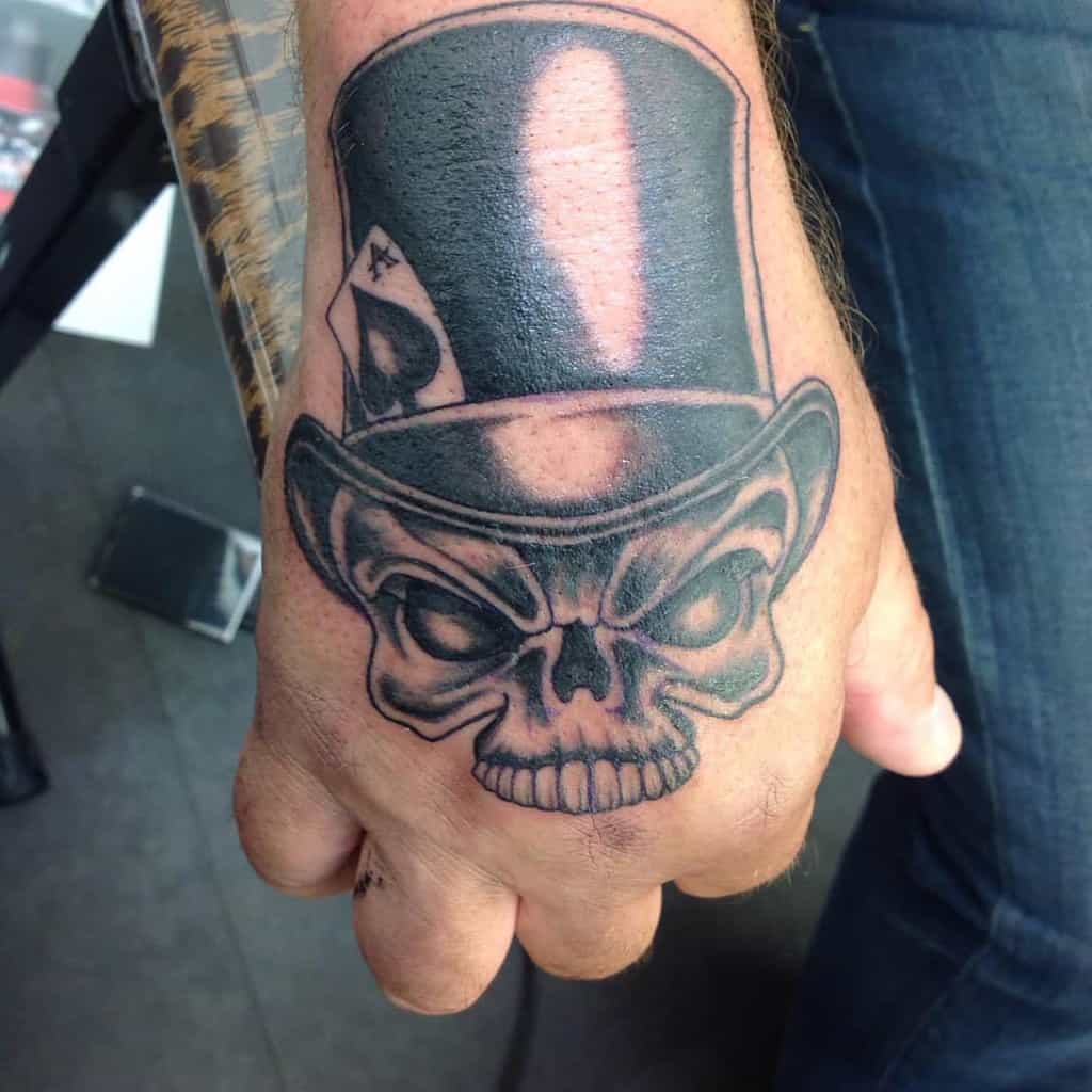 A black-and-white tattoo of a skull wearing a top hat, with an Ace of Spades card tucked into the hatband