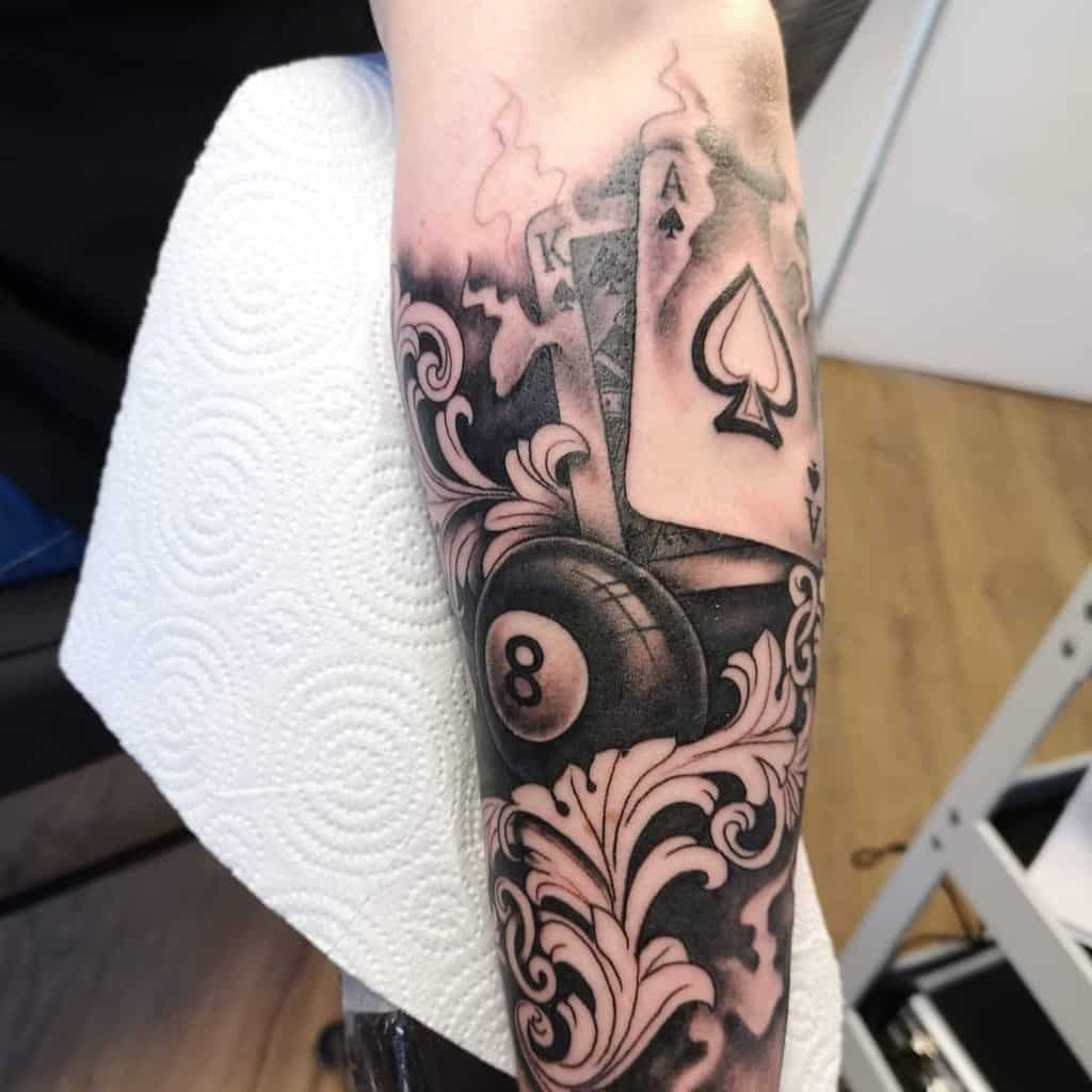 A black-and-white tattoo featuring the Ace and King of Spades cards, smoke rising from them, and an 8-ball with ornate details
