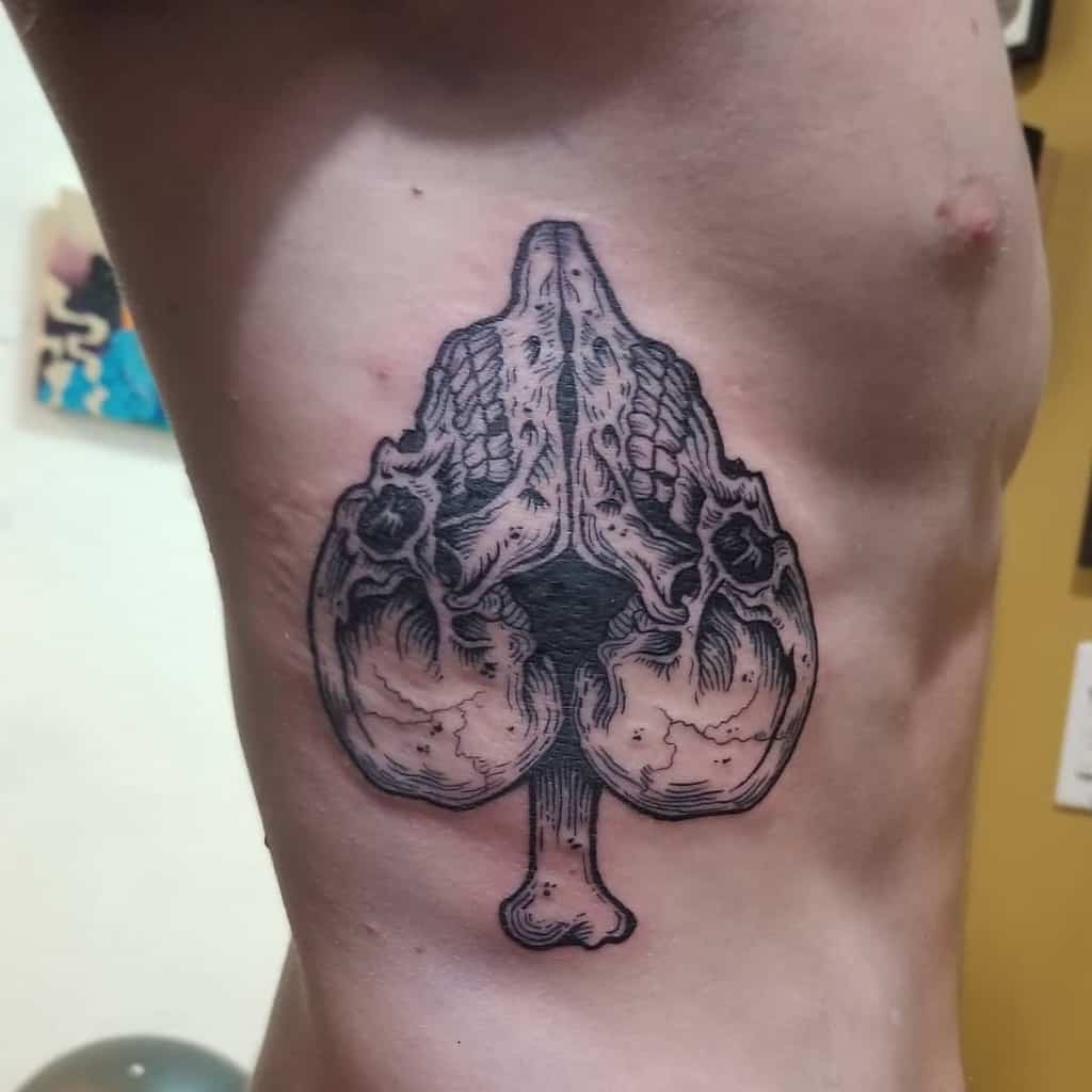 A black-and-white tattoo of a skull and bones, forming the shape of the Ace of Spades symbol