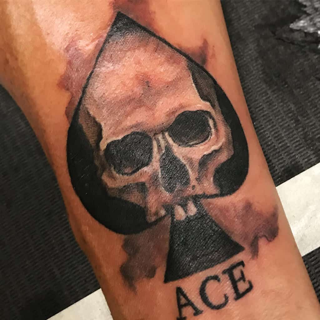 A tattoo of the Ace of Spades with a skull in the center, surrounded by dark shading and a splash of color, with the word "ACE" beneath it