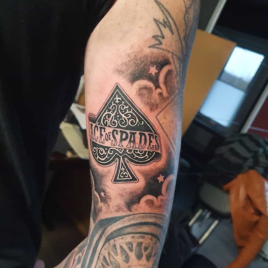 A black-and-white tattoo of the Ace of Spades symbol with ornate detailing, surrounded by clouds and stars, with the text "Ace of Spades" placed across it 