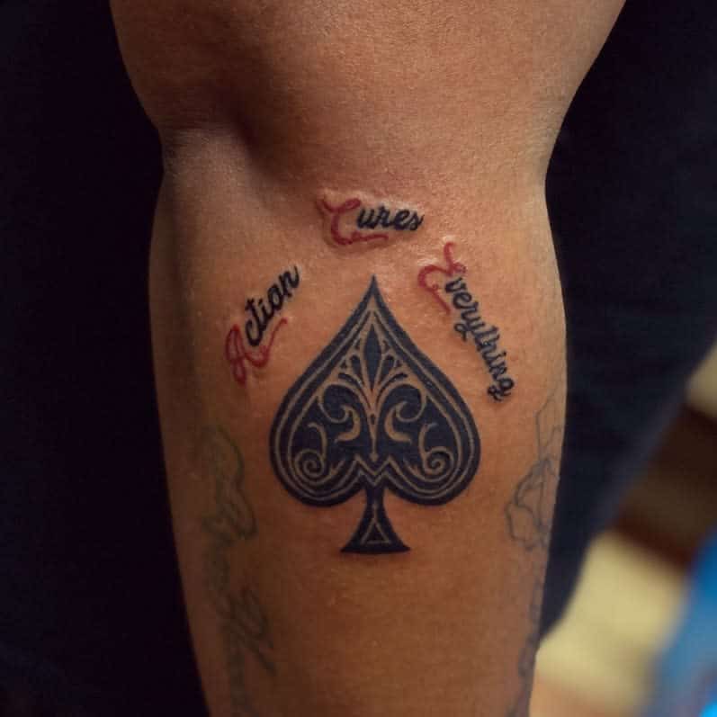 A tattoo featuring an Ace of Spades symbol with ornate detailing, surrounded by the words "Action Cures Everything" in bold lettering