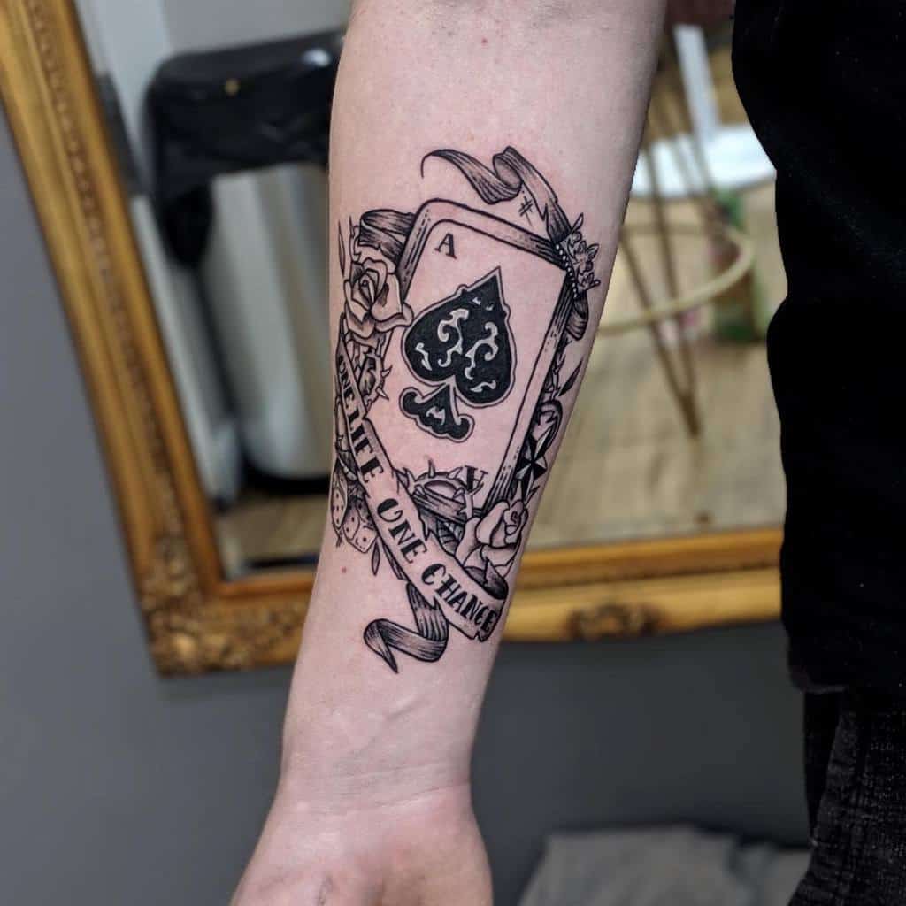 A black-and-white tattoo of the Ace of Spades card surrounded by roses, crown, two dices and ribbon, with the words "One Life One Chance" written across the ribbon
