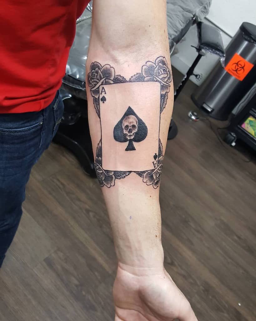 A black-and-white tattoo of the Ace of Spades card with a skull in the center, surrounded by detailed roses