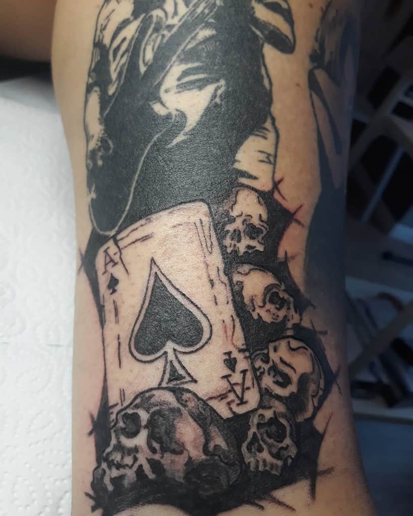 A black-and-white tattoo featuring the Ace of Spades card with several skulls surrounding it