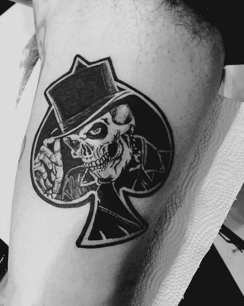 A black-and-white tattoo of a skeleton wearing a top hat, with the skeleton and hat integrated into the shape of the Ace of Spades
