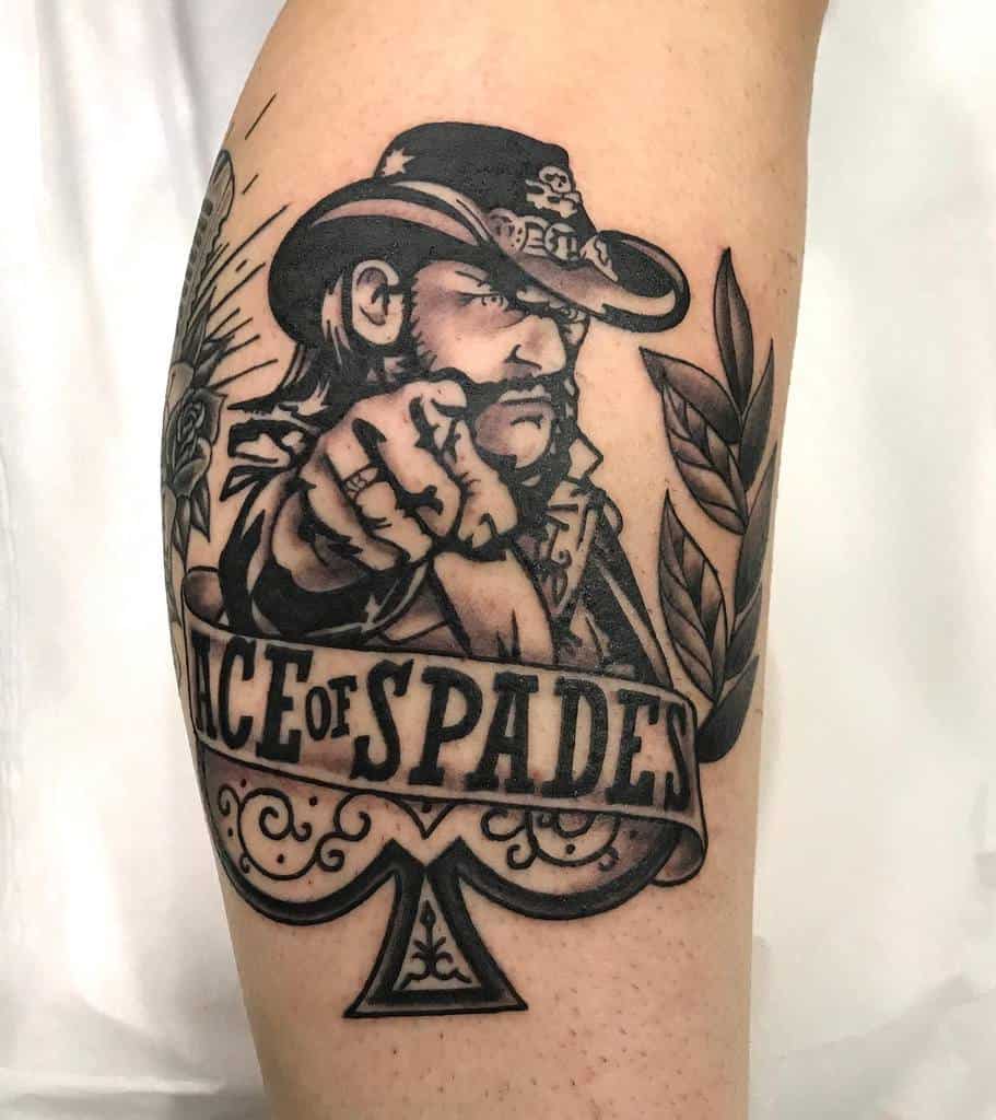 A black-and-white tattoo of a man in a cowboy hat pointing inside a spade design, surrounded by decorative elements like roses and leaves, with "Ace of Spades" written 