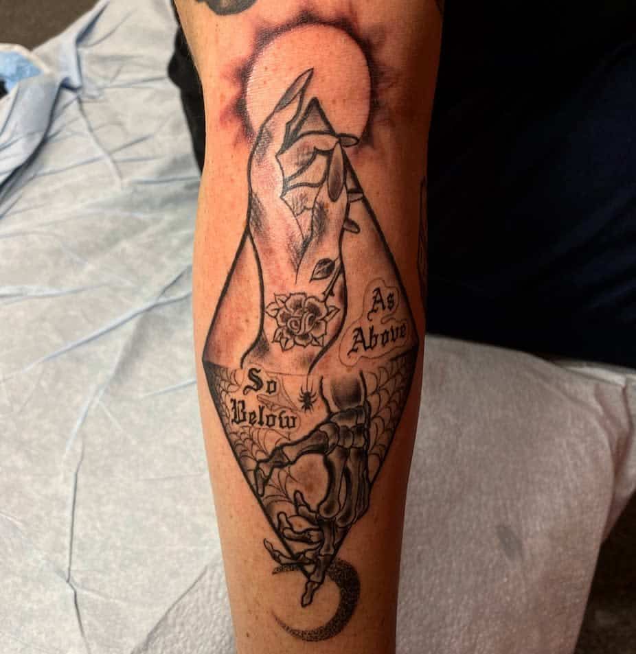 Detailed blackwork tattoo featuring the phrase 'As Above, So Below,' showcasing a mystical hand gesture with intricate elements like a rose, web, skeletal hand, and crescent moon, symbolizing the duality of existence