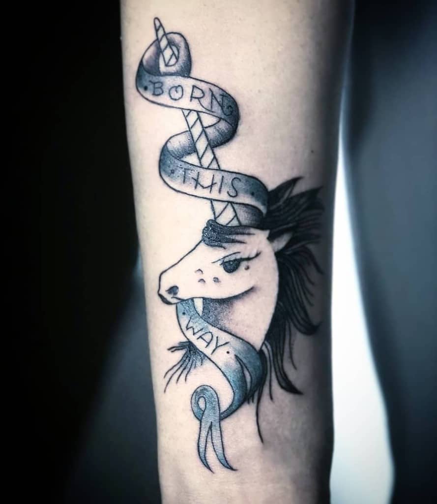 Black and white tattoo of a unicorn with a ribbon saying "Born This Way" on an arm