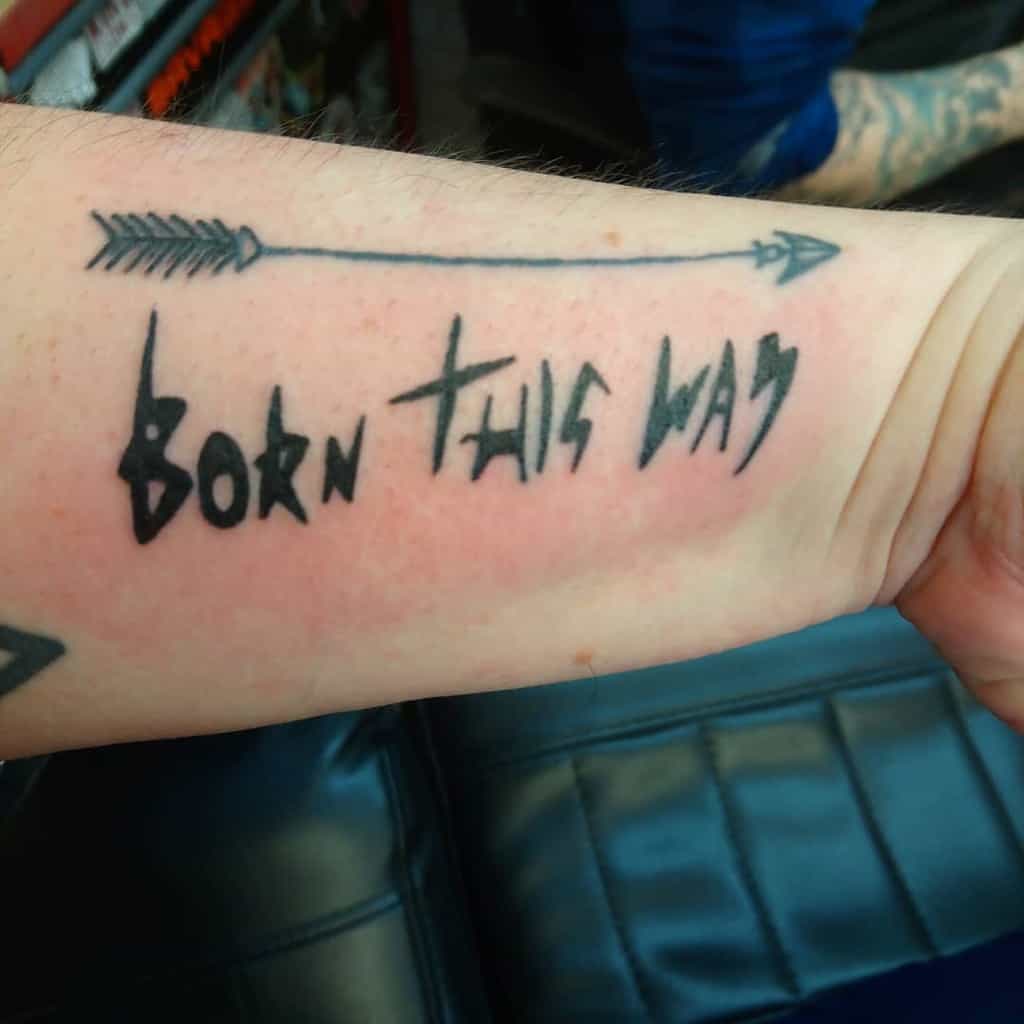 Close-up of a tattoo on an arm with an arrow and the text "Born This Way" in stylized lettering
