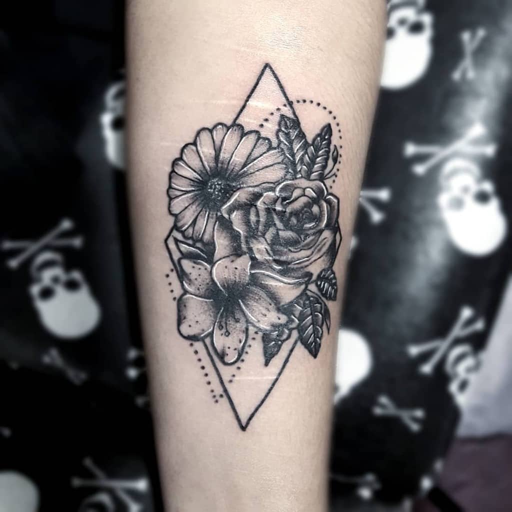 Black and white floral tattoo on forearm, featuring a rose, daisy, and leaves inside a geometric diamond shape