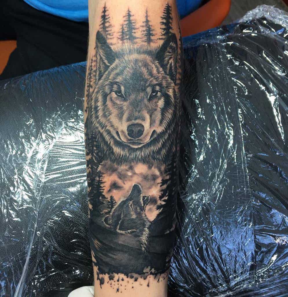 Realistic blackwork tattoo of a wolf on forearm, featuring a large wolf's face with trees in the background and a smaller howling wolf below