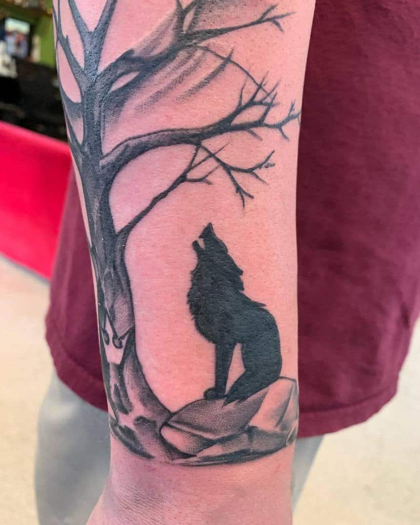 A striking tattoo of a lone wolf howling under a tree with bare branches graces the person's forearm