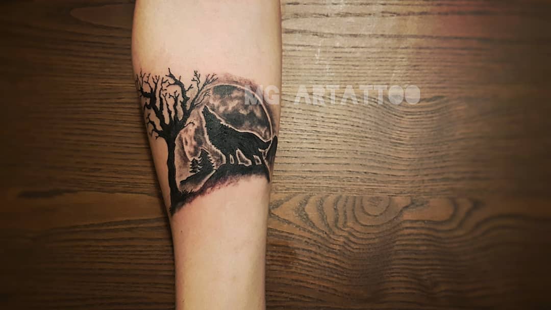 A striking tattoo of a lone wolf howling at a luminous full moon, with silhouetted trees forming a haunting backdrop on a person's arm