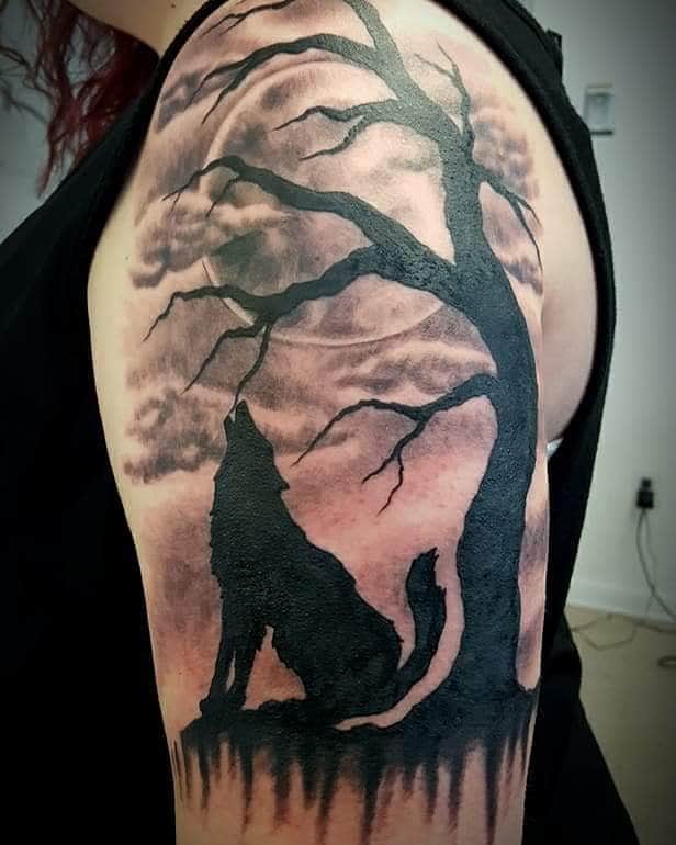 Silhouette blackwork tattoo on upper arm, featuring a howling wolf under a large, barren tree against a cloudy sky background