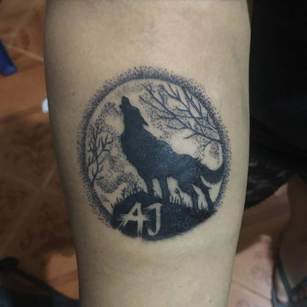 Circular blackwork tattoo with a silhouette of a howling wolf surrounded by trees, and the initials 'AJ' at the bottom