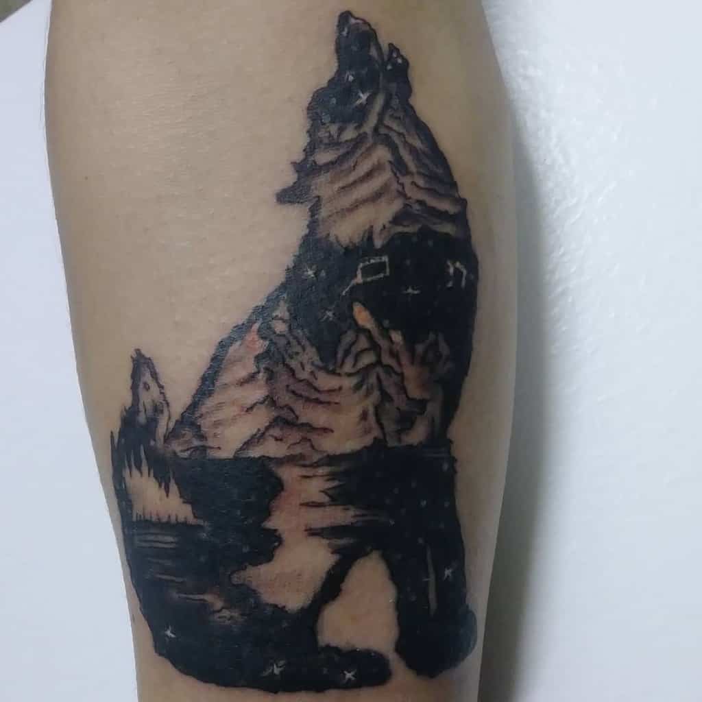 A mesmerizing tattoo of a wolf howling captures the essence of night, with a stunning night scene and majestic mountains seamlessly integrated into the design