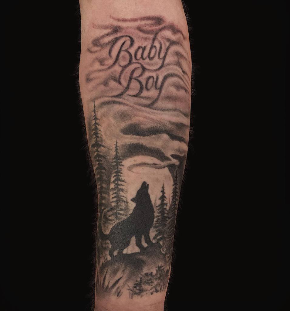 Tattoo of a wolf howling at the moon with trees, accompanied by the "Baby Boy" text on an arm