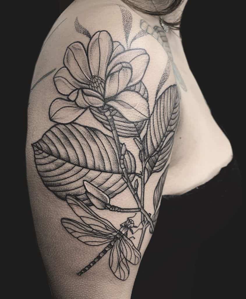 A stunning black and gray tattoo featuring a magnolia intertwined with a graceful dragonfly adorns the person's upper arm