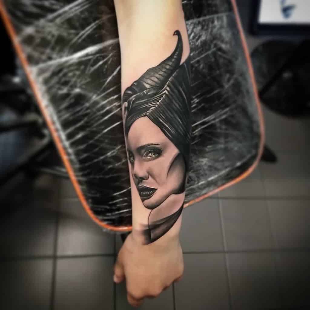 Detailed black-and-gray forearm tattoo of Maleficent with a fierce expression, emphasizing her iconic horns and bold features