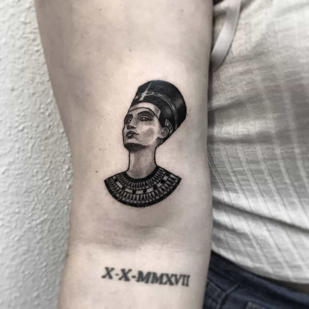 Tattoo of Nefertiti on an arm, next to a Roman numeral date tattoo