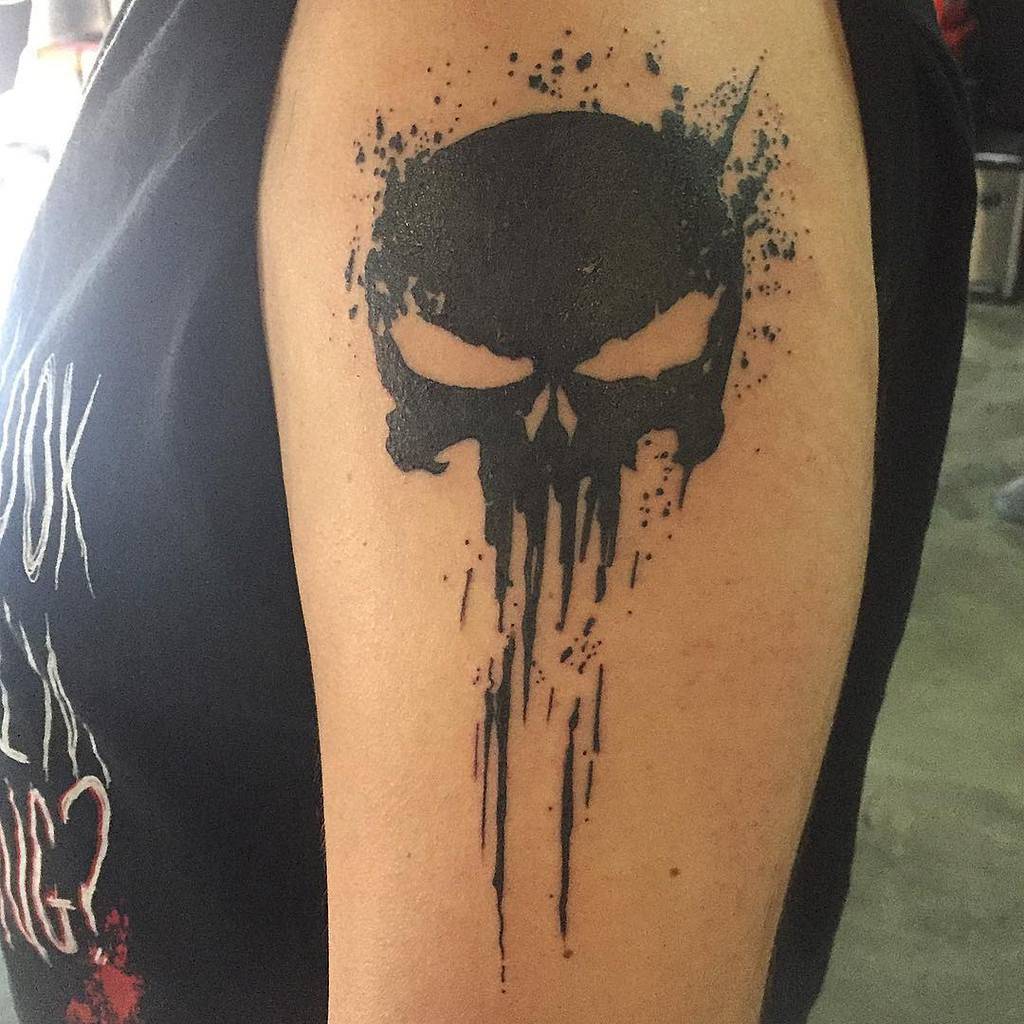 Blackwork Punisher Skull tattoo on the upper arm with splattered ink effects, creating a bold and edgy appearance
