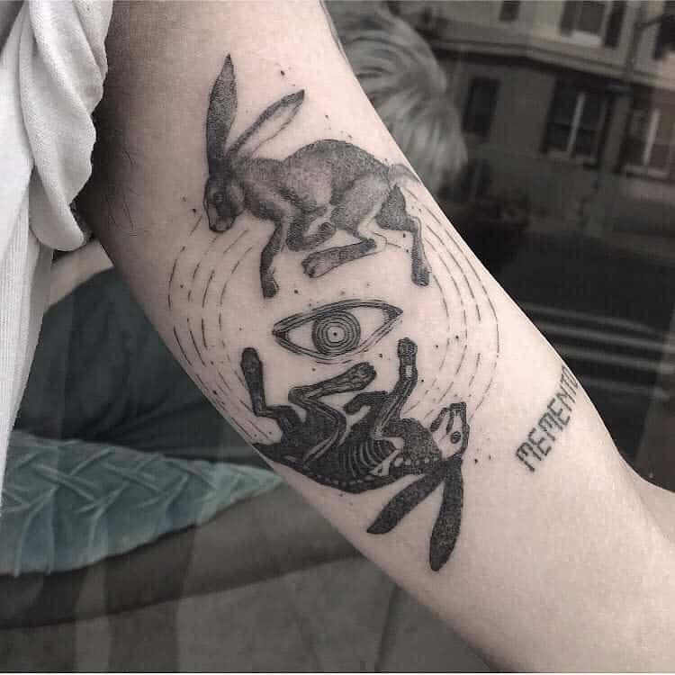 Abstract tattoo featuring two rabbits, one in mid-jump and the other with a skeleton body, surrounded by concentric lines and an eye in the center