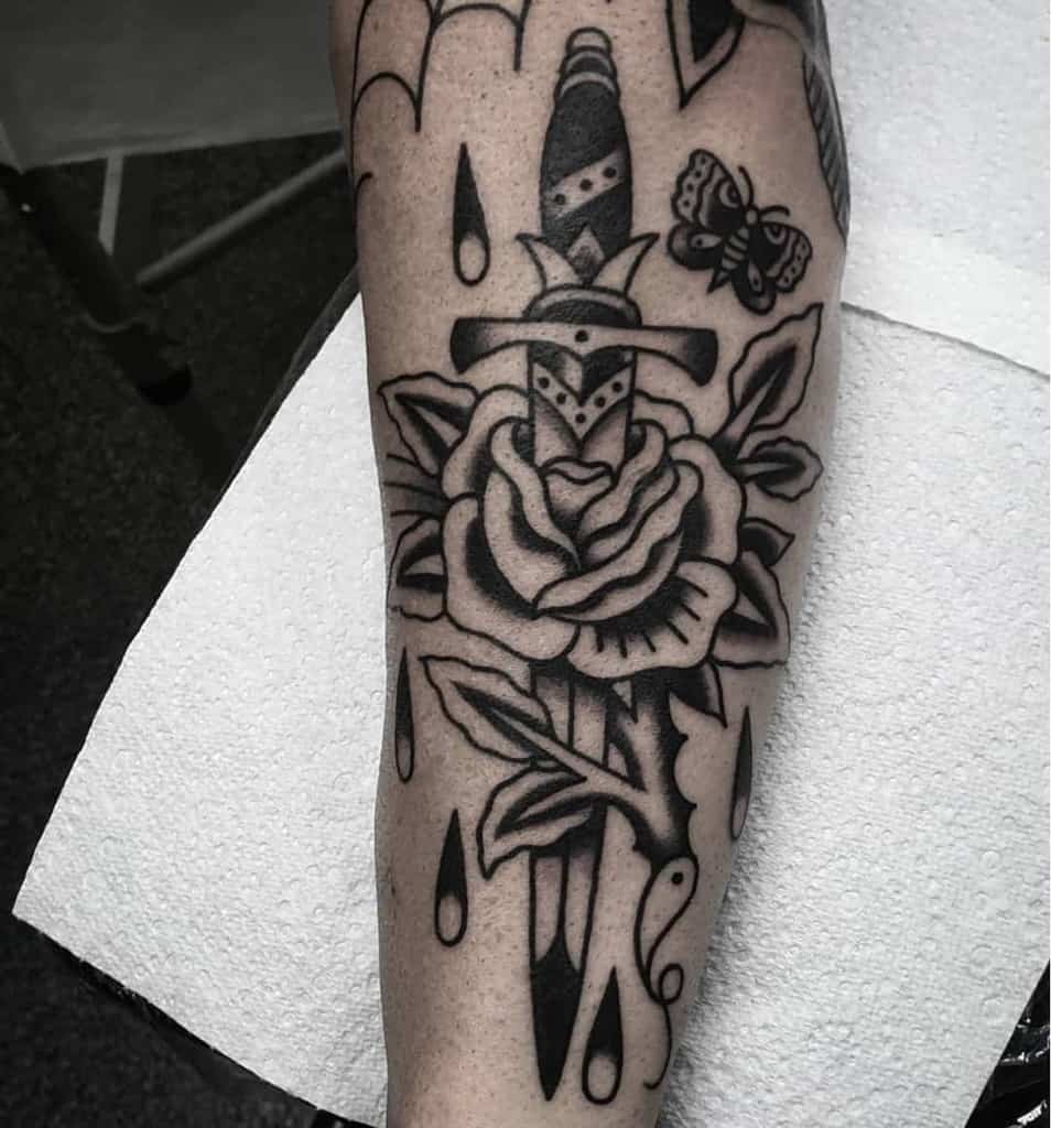 Black ink tattoo of a dagger through a rose with raindrops and a butterfly on a forearm