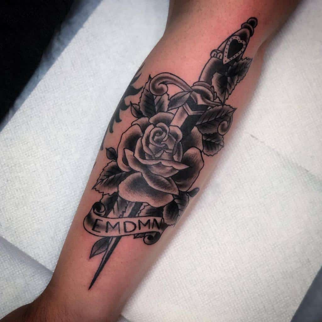 A black and white tattoo of a dagger, rose, and banner with "EMDMN" on an arm