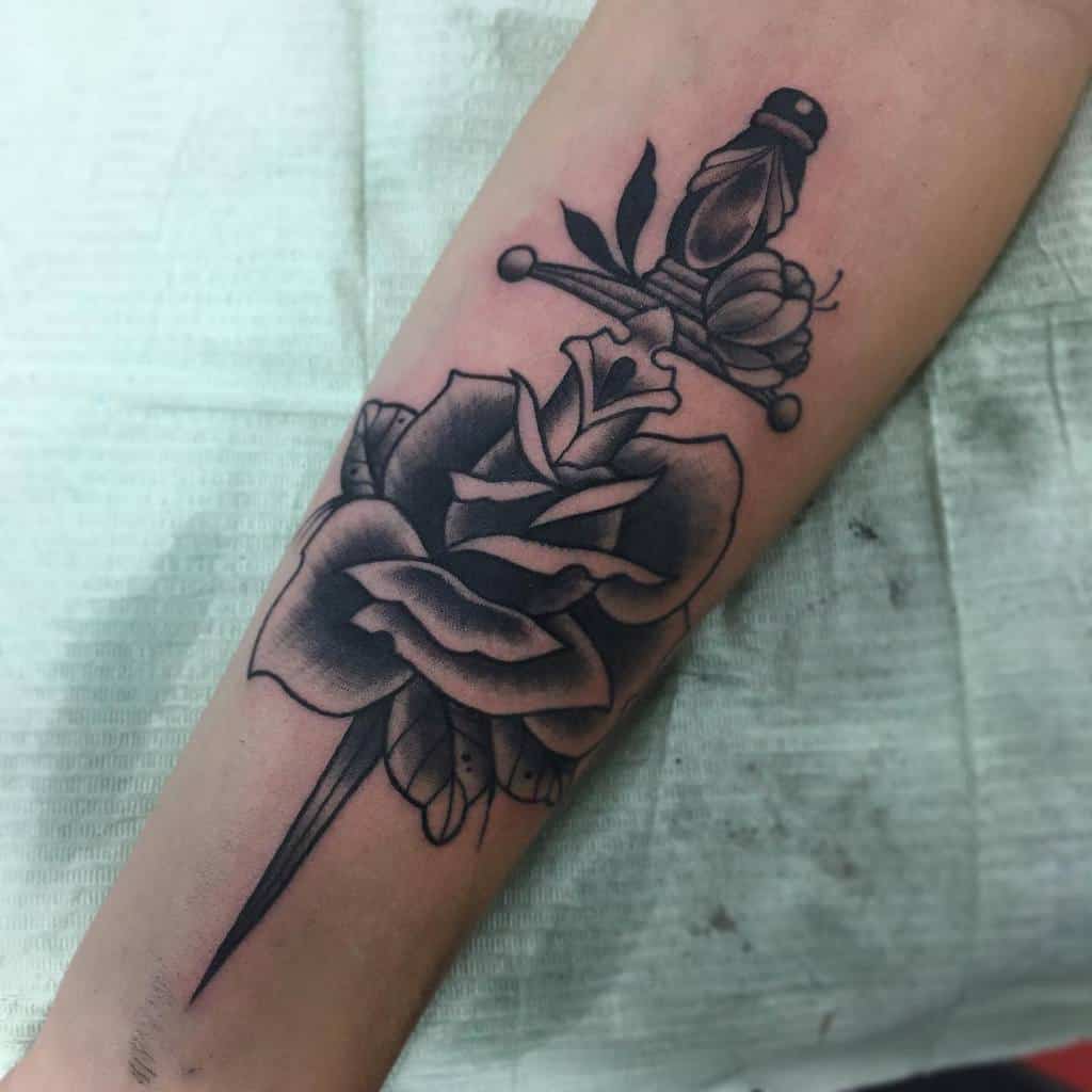 Tattoo of a dagger piercing a rose on a forearm, in black and grey ink