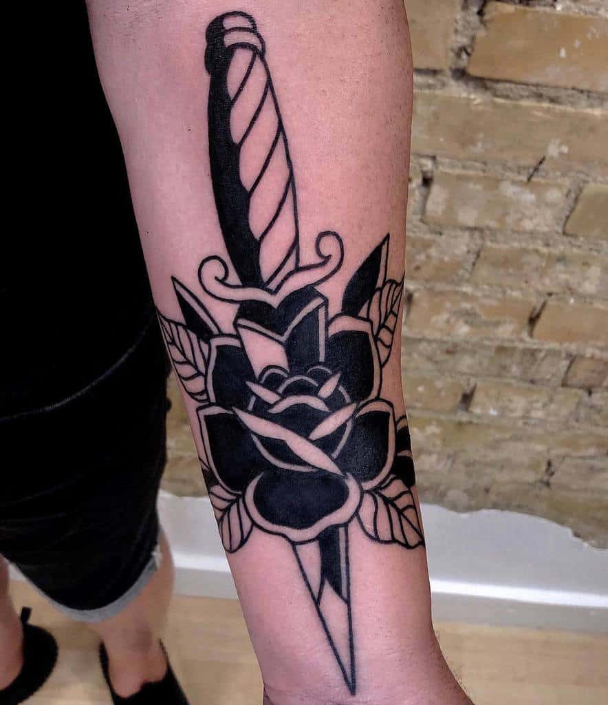 Black tattoo of a dagger through a black rose on a forearm