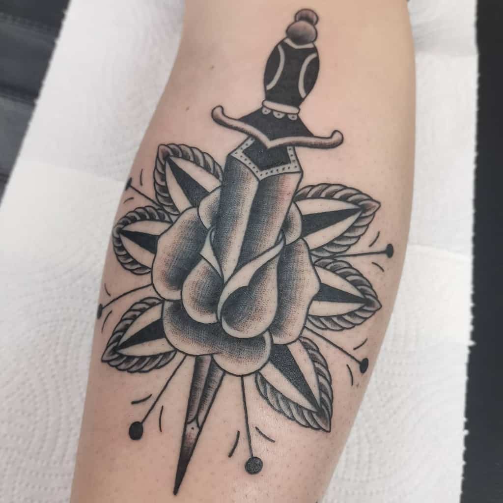 Tattoo of a dagger piercing a rose with detailed shading and leaf accents on the arm