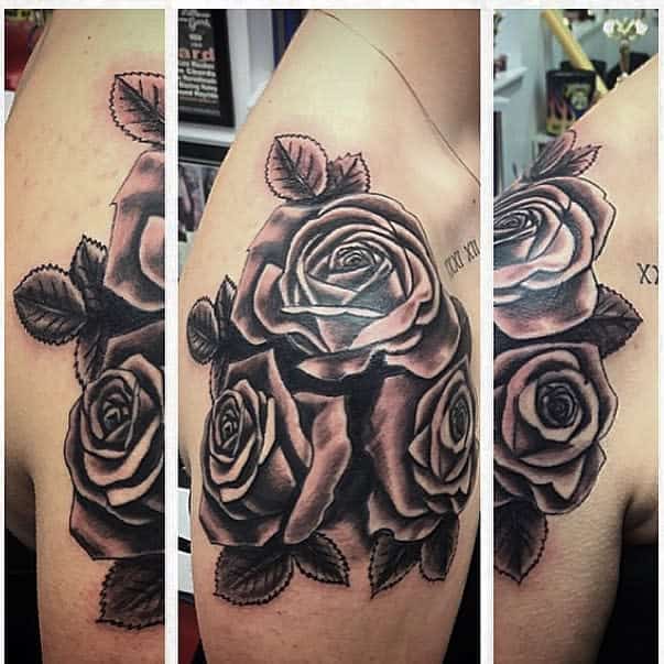 A realistic black and grey rose tattoo on the shoulder, featuring three detailed roses with shading and leaves, accompanied by an inscription