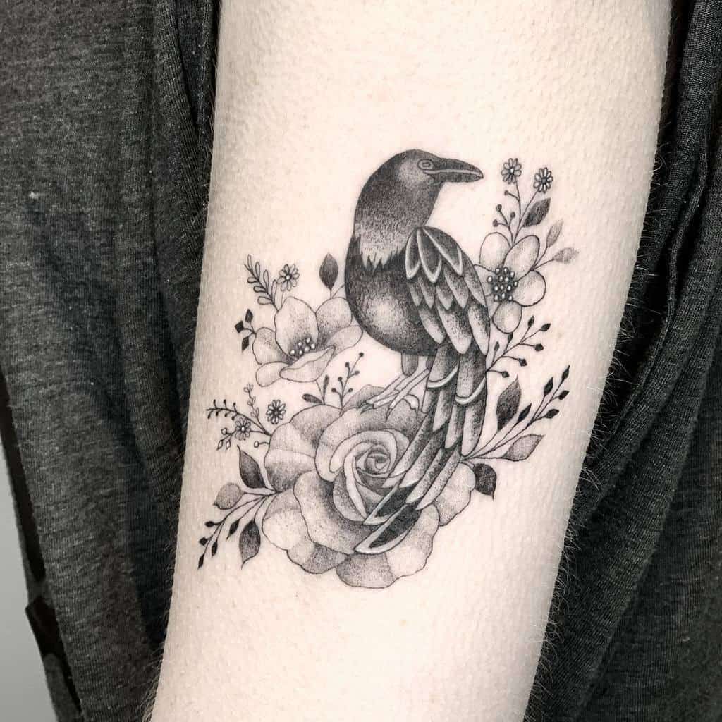 Black and grey tattoo featuring a detailed bird perched on blooming roses, surrounded by delicate floral accents and leaves