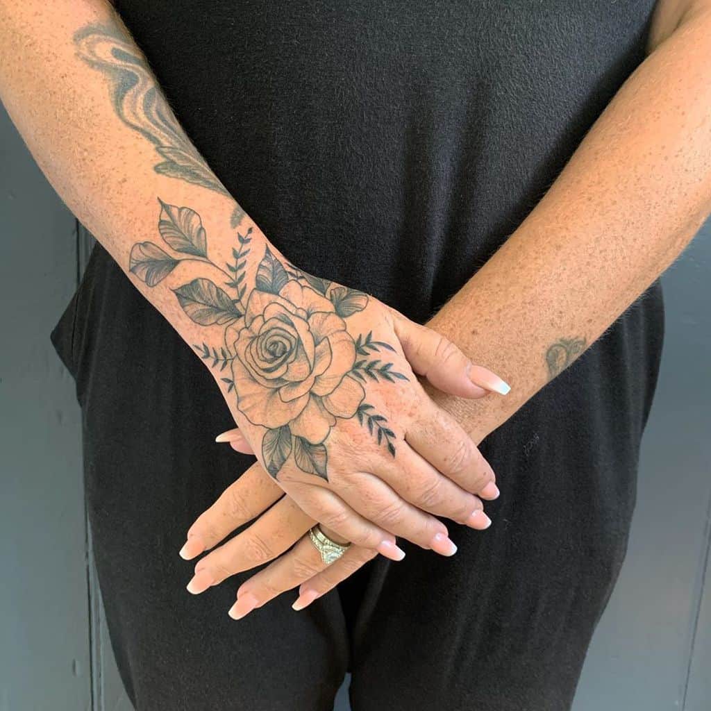 Elegant hand tattoo featuring a beautifully shaded rose as the focal point, surrounded by delicate leaves and subtle vine details extending towards the wrist and forearm