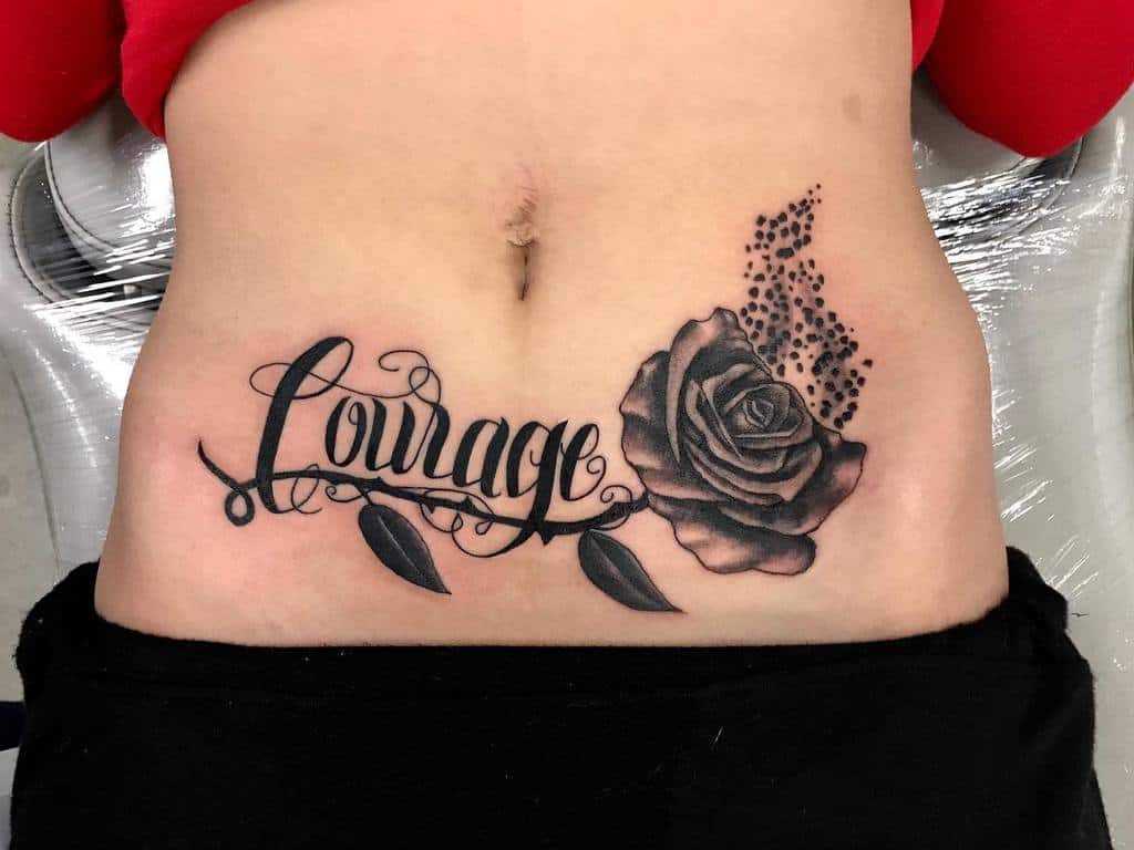 Bold lower abdominal tattoo showcasing the word 'Courage' in elegant script, intertwined with vines and leaves, complemented by a detailed blackwork rose and a scattering dot effect for added depth and texture