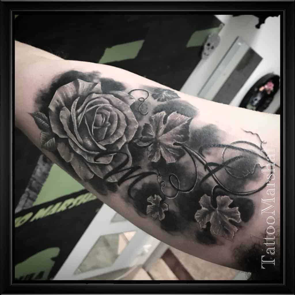 Stunning black and grey tattoo featuring a meticulously shaded rose at the center, surrounded by detailed vine-like leaves and swirling accents, creating a harmonious balance of depth and artistry