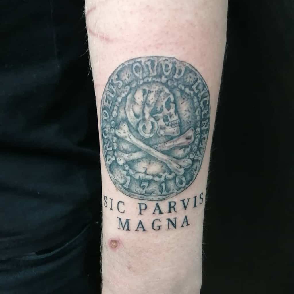 Tattoo of a coin featuring a skull and crossbones with the text "Sic Parvis Magna" underneath